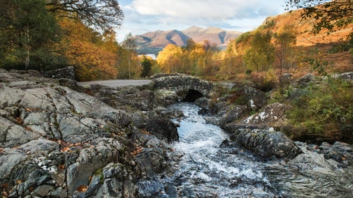 A stone bridge over a fast-flowing river flanked on either side with hard, grey stones and autumnal-leaved trees. Is the distance you can see warm evening light on dramatic-looking hills.